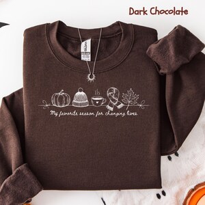 May include: Dark chocolate-colored sweatshirt with a crew neck and long sleeves. The front features a white line drawing of fall-themed icons: a pumpkin, hat, coffee cup, scarf, and leaf. The text "My favorite season for changing lives" is below the icons.