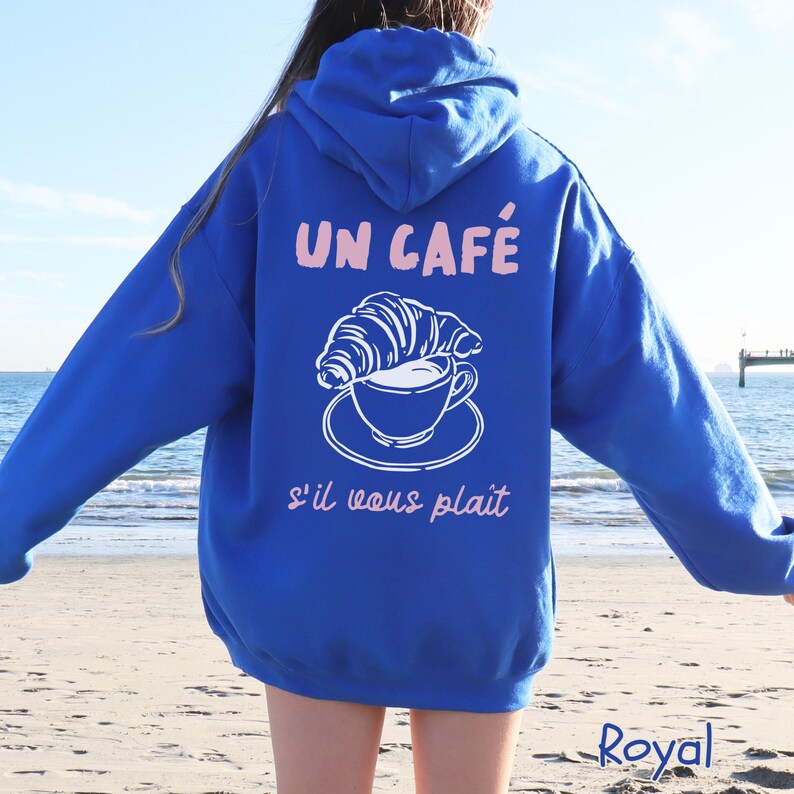 May include: A royal blue hooded sweatshirt featuring a white line art graphic of a croissant resting on a coffee cup and saucer. The text "UN CAF&Eacute;" appears in light pink above the graphic, and "s'il vous pla&icirc;t" is printed in light pink below.