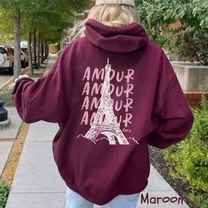 K&ouml;nnte beinhalten: Ein kastanienbrauner Kapuzenpullover mit einer wei&szlig;en Grafik auf der R&uuml;ckseite. Das Design zeigt eine Strichzeichnung des Eiffelturms, zentriert zwischen vier &uuml;bereinanderliegenden Zeilen des Wortes AMOUR in stilisierter Schrift, mit Paris darunter.