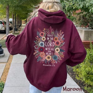 Puede incluir: Sudadera con capucha color granate con un dise&ntilde;o floral y el texto "Trust in the Lord with all your heart" de Proverbios 3:5. El dise&ntilde;o presenta flores y hojas coloridas. La sudadera tiene capucha y mangas largas.