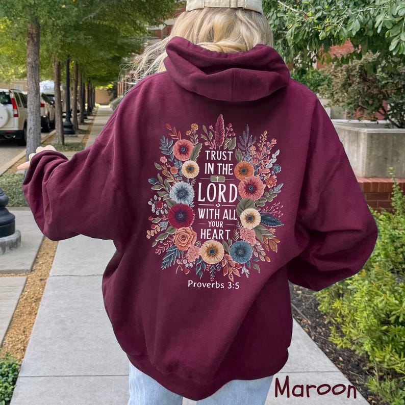 Puede incluir: Sudadera con capucha color granate con un dise&ntilde;o floral y el texto "Trust in the Lord with all your heart" de Proverbios 3:5. El dise&ntilde;o presenta flores y hojas coloridas. La sudadera tiene capucha y mangas largas.