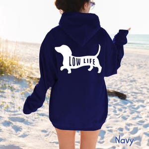 以下が含まれることがあります： 白いダックスフントのシルエットと犬の体内に「LOW LIFE」の文字がプリントされたネイビーブルーのパーカー。 パーカーはビーチで人に着られています。