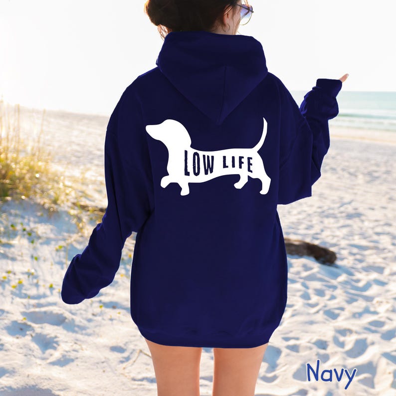 以下が含まれることがあります： 白いダックスフントのシルエットと犬の体内に「LOW LIFE」の文字がプリントされたネイビーブルーのパーカー。 パーカーはビーチで人に着られています。