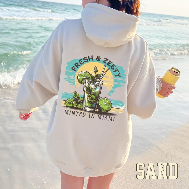 Turn up the tropical energy with our Fresh & Zesty – Minted in Miami hoodie, featuring a bold mojito graphic set against a dreamy Miami sunset beach scene. This eye-catching back print channels sunny coastal vibes, citrusy freshness, and laid-back luxury—perfect for sipping mojitos or dreaming of South Beach. A go-to for travel, lounging, or gifting anyone who loves cocktails, sunshine, and Miami heat.  Ideal gift for cocktail lovers, beachgoers, or anyone with a thing for Miami vibes.