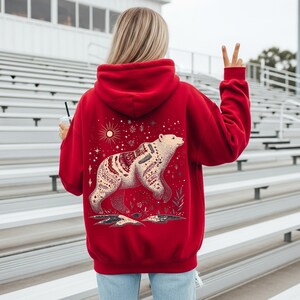May include: A vibrant red hoodie with a detailed bear illustration. The bear is cream-coloured with a patterned design. The hoodie has a drawstring hood and long sleeves. The design includes a sun, stars, and floral elements. The hoodie is a comfortable garment.