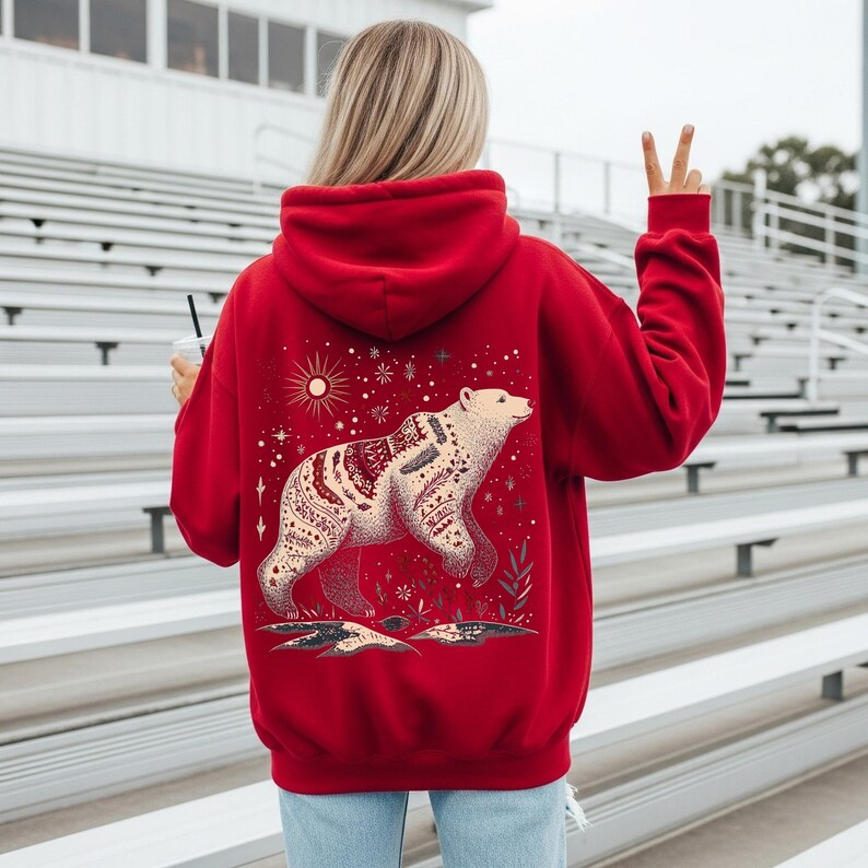 May include: A vibrant red hoodie with a detailed bear illustration. The bear is cream-coloured with a patterned design. The hoodie has a drawstring hood and long sleeves. The design includes a sun, stars, and floral elements. The hoodie is a comfortable garment.