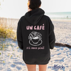 May include: A black hooded sweatshirt featuring a white line art graphic of a croissant resting on a coffee cup. The text "UN CAF&Eacute;" appears in light pink above the graphic, and "s'il vous pla&icirc;t" is printed in light pink below the illustration.