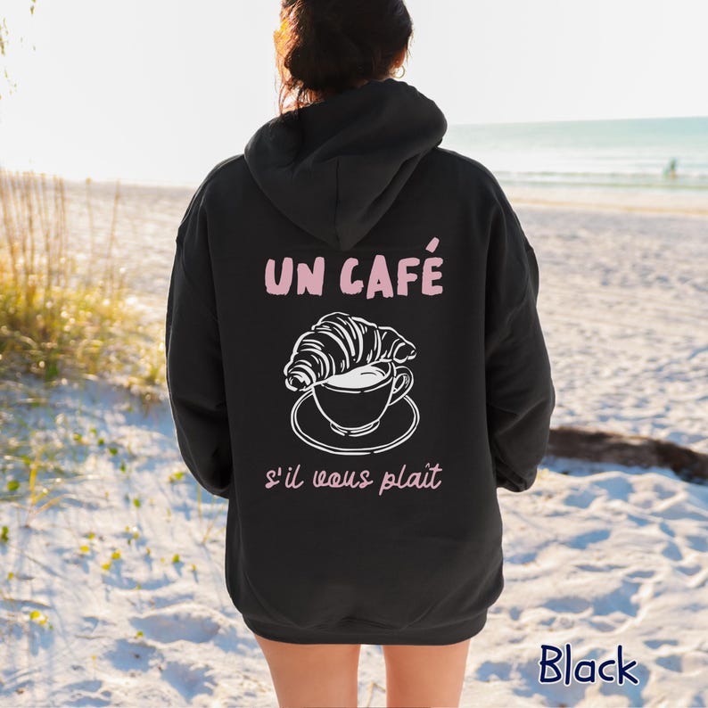 May include: A black hooded sweatshirt featuring a white line art graphic of a croissant resting on a coffee cup. The text "UN CAF&Eacute;" appears in light pink above the graphic, and "s'il vous pla&icirc;t" is printed in light pink below the illustration.