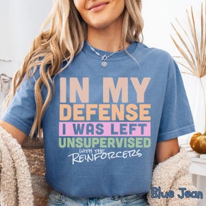May include: Blue jean-coloured t-shirt with the text "IN MY DEFENSE I WAS LEFT UNSUPERVISED WITH THE REINFORCERS" in various colours. The shirt has a crew neck and short sleeves. The text "Blue Jean" is at the bottom right.