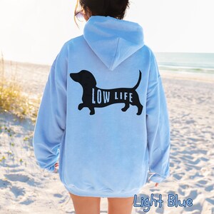 以下が含まれることがあります： 黒いダックスフンドのデザインと「LOW LIFE」の文字がプリントされた、ライトブルーのフード付きスウェットシャツ。フードと長袖が付いています。「Light Blue」の文字が画像の下部に印刷されています。