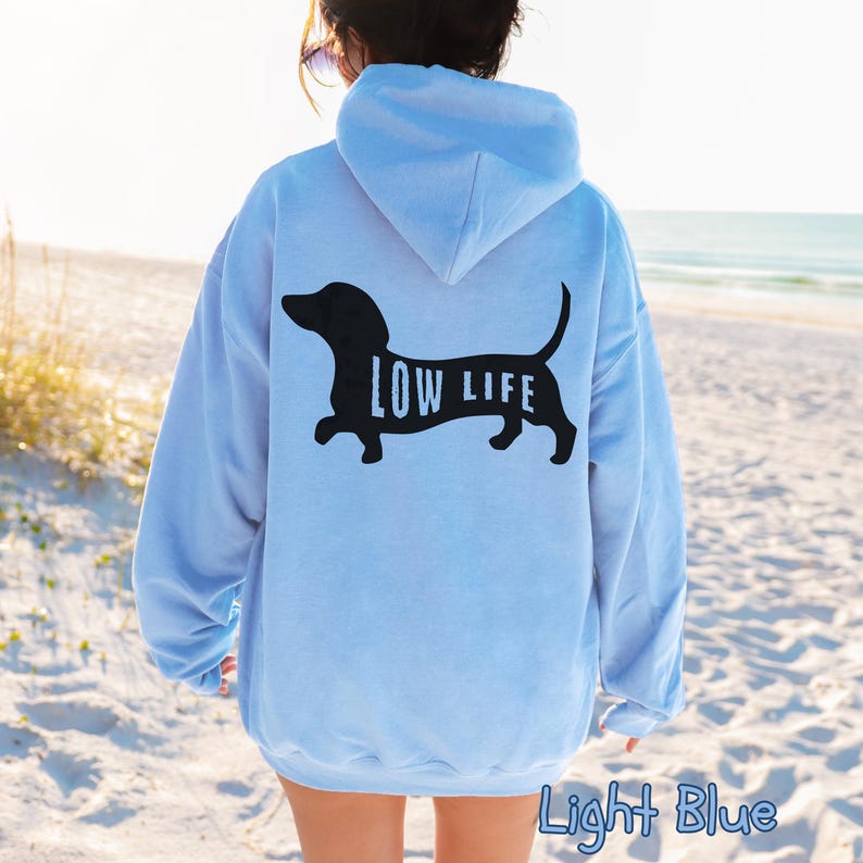 以下が含まれることがあります： 黒いダックスフンドのデザインと「LOW LIFE」の文字がプリントされた、ライトブルーのフード付きスウェットシャツ。フードと長袖が付いています。「Light Blue」の文字が画像の下部に印刷されています。