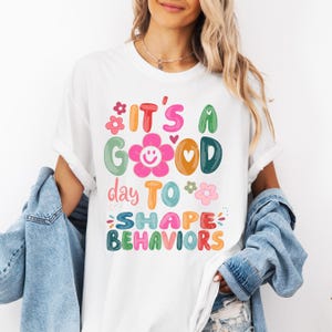 May include: White t-shirt with a colourful graphic that reads "It's a Good Day to Shape Behaviors." The design features flowers, hearts, and a smiling flower. The shirt has short sleeves and a relaxed fit.