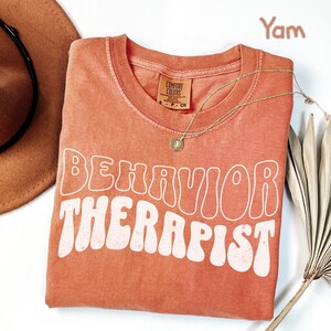 May include: A coral-colored t-shirt with the words "BEHAVIOR THERAPIST" in white, retro-style lettering. A gold necklace with a pendant rests on the shirt. The shirt is folded and placed on a white surface, with a brown hat and dried plant in the background.