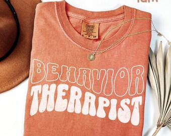 May include: A coral-colored t-shirt with the words "BEHAVIOR THERAPIST" in white, retro-style lettering. A gold necklace with a pendant rests on the shirt. The shirt is folded and placed on a white surface, with a brown hat and dried plant in the background.