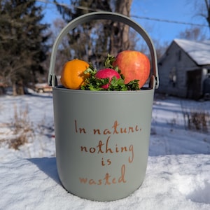 May include: A grey bucket with a handle, containing an orange, a red apple, a radish, and green herbs. The bucket has the text "In nature, nothing is wasted" in copper-coloured script. The bucket is in the snow.