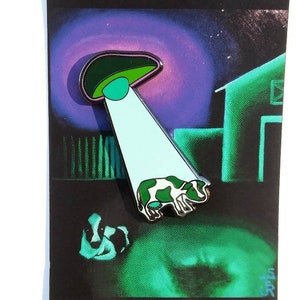 Könnte beinhalten: Eine grüne und violette Emaille-Anstecknadel mit einer Cartoon-Kuh, die von einem UFO entführt wird. Die Anstecknadel trägt den Text "JESSEJFR" und "MISSING TIME" auf der Karte.