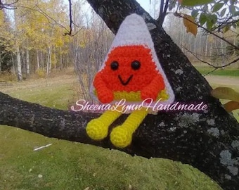 Pode incluir: Um personagem de milho doce de crochê com um rosto sorridente, corpo laranja e amarelo e parte superior branca. O personagem está sentado em um galho de árvore com folhagem de outono no fundo.