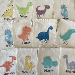 Puede incluir: Diez peque&ntilde;as bolsas de tela blanca con cierre de cord&oacute;n. Cada bolsa presenta un dise&ntilde;o de dinosaurio diferente y un nombre impreso debajo del dinosaurio. Los nombres son: Ashli, Noah, Mimi, Ethan, Kira, Ali, Haroon, Nicolette, Maggie y Andrew.