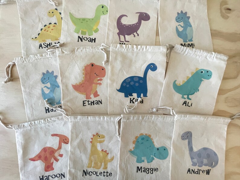 Puede incluir: Diez peque&ntilde;as bolsas de tela blanca con cierre de cord&oacute;n. Cada bolsa presenta un dise&ntilde;o de dinosaurio diferente y un nombre impreso debajo del dinosaurio. Los nombres son: Ashli, Noah, Mimi, Ethan, Kira, Ali, Haroon, Nicolette, Maggie y Andrew.