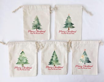 Puede incluir: Cinco bolsas de algodón blanco con cordón de ajuste, decoradas con un diseño de acuarela de un árbol de Navidad verde con una estrella roja en la parte superior. Cada bolsa tiene el texto "Merry Christmas! From: The Mercado Family" impreso en ella.