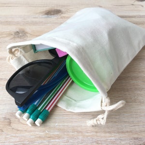 Puede incluir: Una bolsa de tela blanca con un recipiente verde con tapa, gafas de sol y l&aacute;pices en su interior.