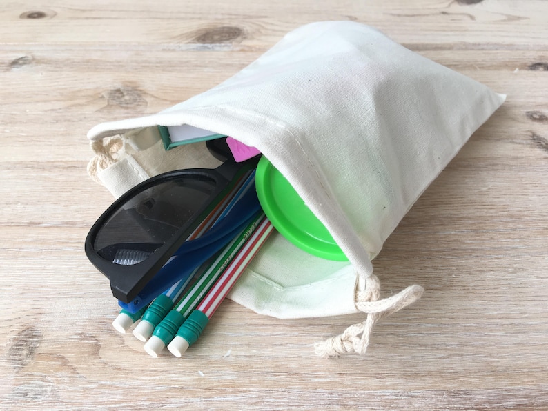 Puede incluir: Una bolsa de tela blanca con un recipiente verde con tapa, gafas de sol y l&aacute;pices en su interior.