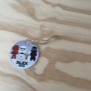 May include: A round, light blue ornament with a string, featuring cartoon characters from Star Wars. The ornament has three characters, a red-haired figure, a Stormtrooper, and a black-clad figure. The text "ALEX 2022" is printed below the characters.