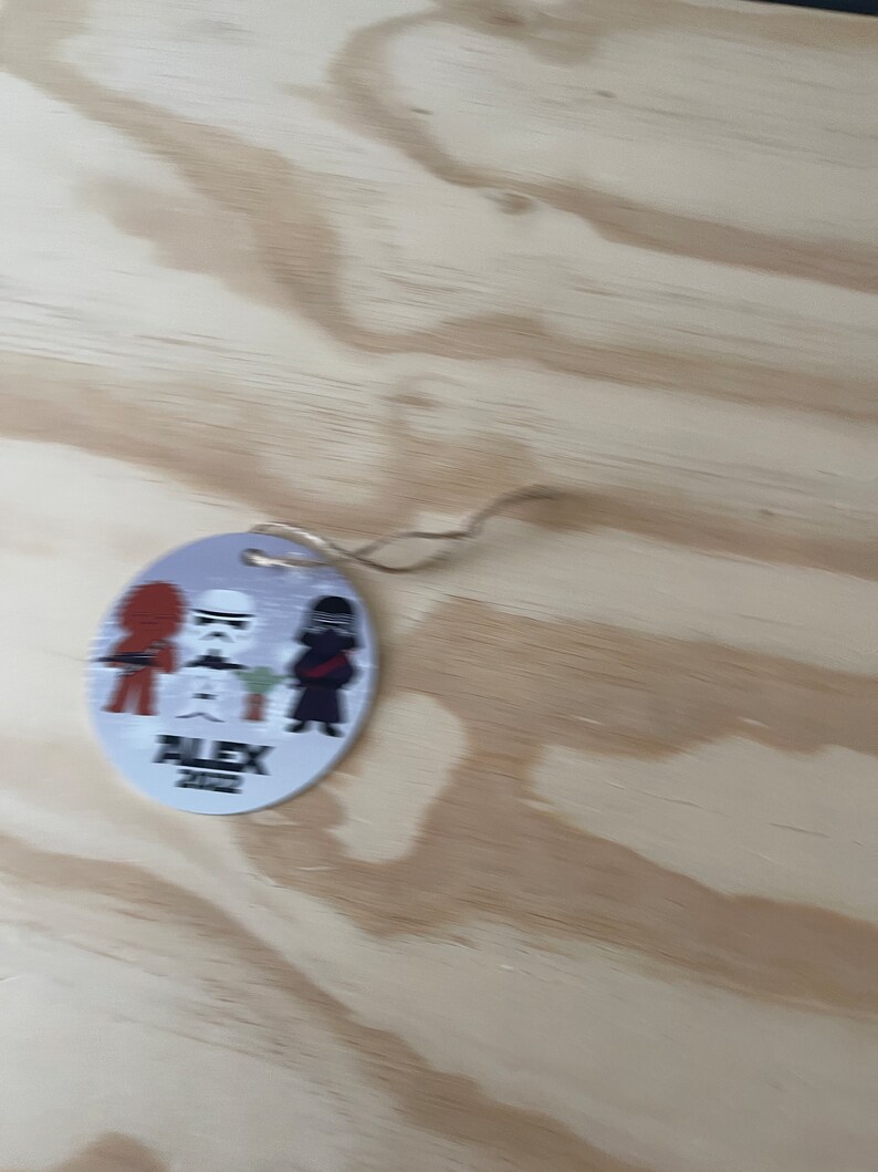 May include: A round, light blue ornament with a string, featuring cartoon characters from Star Wars. The ornament has three characters, a red-haired figure, a Stormtrooper, and a black-clad figure. The text "ALEX 2022" is printed below the characters.