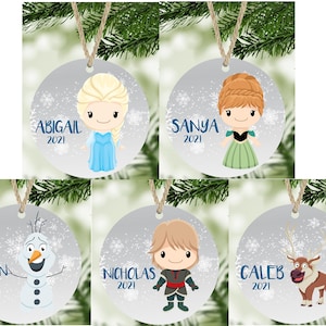 May include: Five round Christmas ornaments with a silver background and a white snowflake pattern. Each ornament features a different character from the movie Frozen. The ornaments are personalized with names and the year 2021. The characters include Elsa, Anna, Olaf, Kristoff, and Sven.