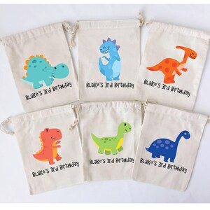 Puede incluir: Seis peque&ntilde;as bolsas de lona blancas con cierre de cord&oacute;n. Cada bolsa presenta una ilustraci&oacute;n de dinosaurio de colores diferente y el texto "Blake's 3rd Birthday".