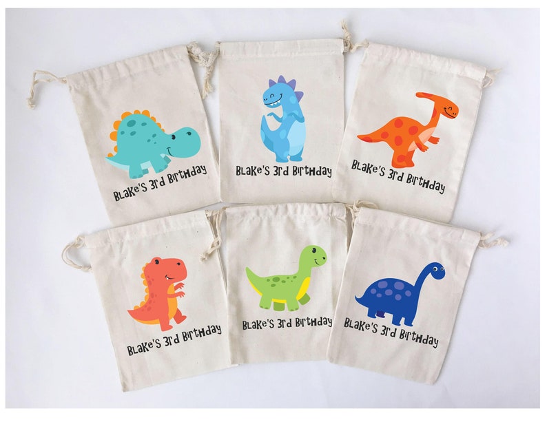 Puede incluir: Seis peque&ntilde;as bolsas de lona blancas con cierre de cord&oacute;n. Cada bolsa presenta una ilustraci&oacute;n de dinosaurio de colores diferente y el texto "Blake's 3rd Birthday".