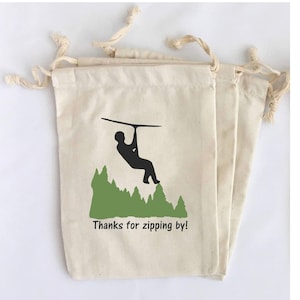 May include: Off-white drawstring bags with a black silhouette of a person ziplining over green trees. The text "Thanks for zipping by!" is printed below the graphic. The bags are made of a natural fabric.