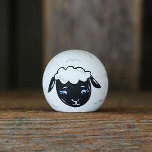May include: A spherical white wooden object featuring a hand-painted black sheep face with curly white wool detail on the forehead, blue eyes, and black ears. The surface of the sphere is decorated with faint, light gray outlines of clouds.