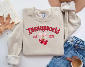 Może przedstawiać: Beżowa bluza z napisem "Disneyworld est 1971" w czerwonych i czarnych literach. Różowa kokardka z dwoma czerwonymi wiśniami zwisa pod napisem.