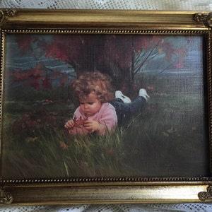May include: A framed print featuring a child in a pink shirt and jeans, lying in grass and holding a leaf. The print is set in a gold-colored ornate frame, with a background of a tree and red leaves.