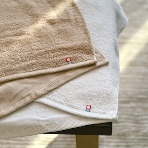 May include: Two towels, one beige and one white, are folded on top of each other. Both towels have a small red circle with a white stripe design in the bottom right corner.