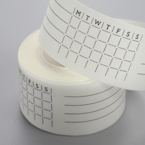 May include: A roll of white adhesive tape featuring a repeating black grid pattern. The design includes the letters M, T, W, T, F, S, S across the top of the grid, representing the days of the week, alongside a section with four horizontal lines.