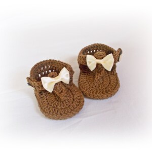 May include: A pair of brown crochet baby shoes featuring a textured knit pattern, a rounded toe, and a small cream-colored satin ribbon bow attached to the front of each shoe. The shoes have a strap design with a small button closure on the side.