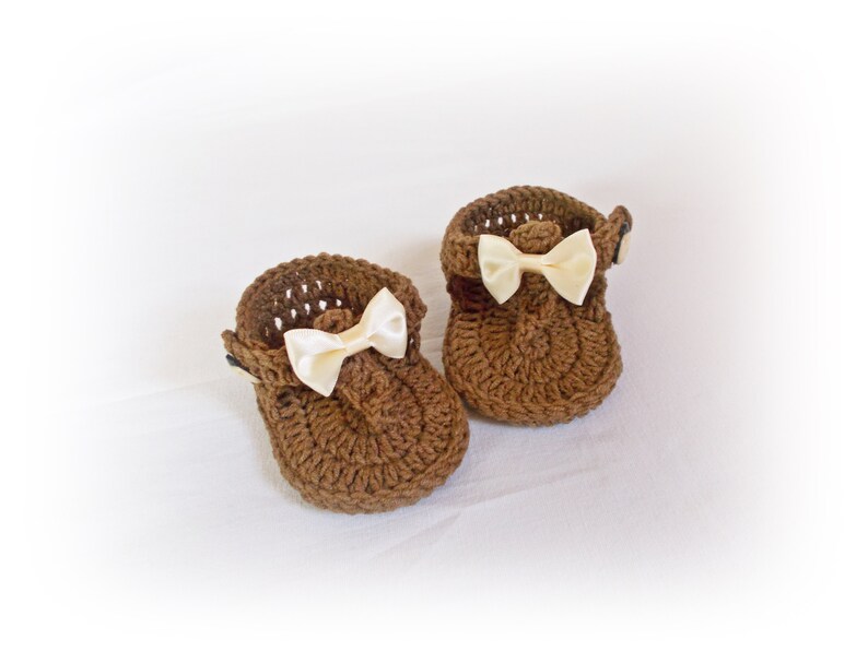 May include: A pair of brown crochet baby shoes featuring a textured knit pattern, a rounded toe, and a small cream-colored satin ribbon bow attached to the front of each shoe. The shoes have a strap design with a small button closure on the side.