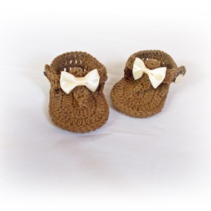 May include: A pair of brown crocheted baby sandals featuring a textured yarn construction, a small cream-colored satin ribbon bow on the front of each shoe, and a side button closure to secure the strap around the ankle area of the footwear.