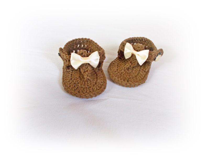 May include: A pair of brown crocheted baby sandals featuring a textured yarn construction, a small cream-colored satin ribbon bow on the front of each shoe, and a side button closure to secure the strap around the ankle area of the footwear.
