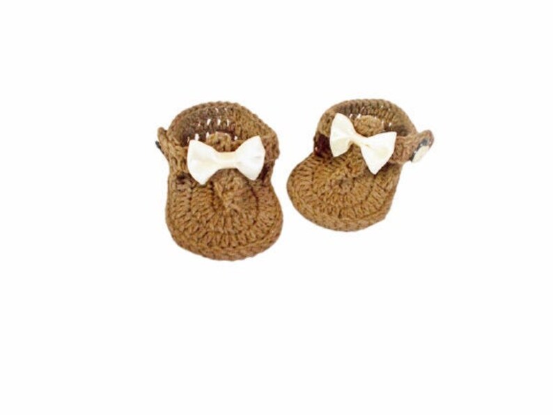 May include: A pair of brown crocheted baby booties featuring a textured yarn construction and a small, off-white satin bow attached to the front of each shoe. The booties have a rounded toe design and a strap detail on the side of each piece.