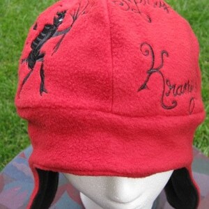 May include: Red fleece hat with black embroidered designs. The designs include a stick figure holding a stick, the word "for us", and the word "remarkable". The hat has black ear flaps.