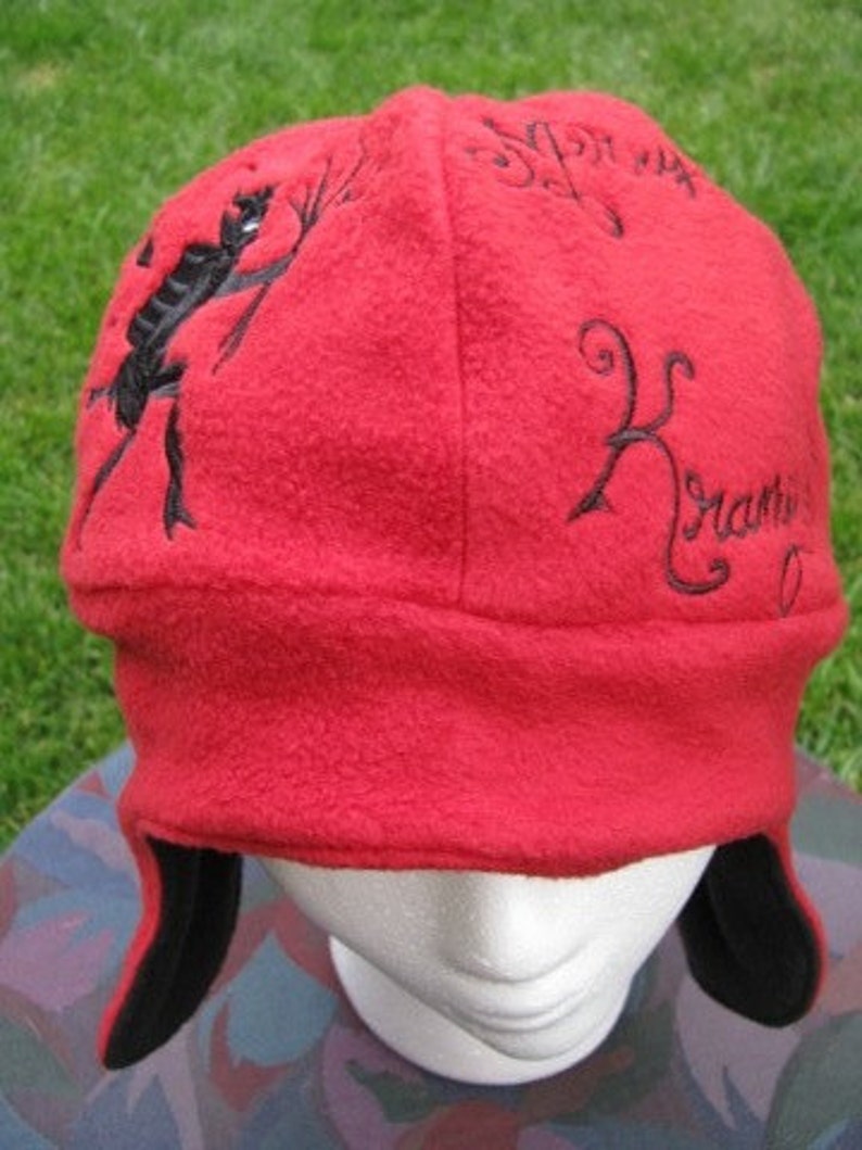 May include: Red fleece hat with black embroidered designs. The designs include a stick figure holding a stick, the word "for us", and the word "remarkable". The hat has black ear flaps.