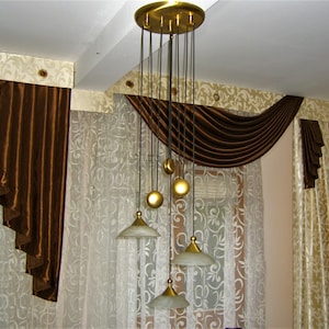 Könnte beinhalten: Ein brauner und goldener Vorhang mit einer dekorativen Schlaufenschal. Die Vorhänge sind an einer weißen Decke mit einem goldenen Kronleuchter mit mehreren Lichtern aufgehängt.