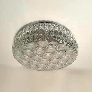 Può includere: Un lampadario da soffitto in vetro trasparente con un design strutturato a forma di bolle. L'apparecchio è rotondo e ha una base in metallo scuro. La luce è montata su un soffitto bianco.
