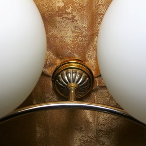 May include: A close-up of a gold and silver ceiling light fixture with two white glass globes. The fixture has a curved arm connecting the two globes.