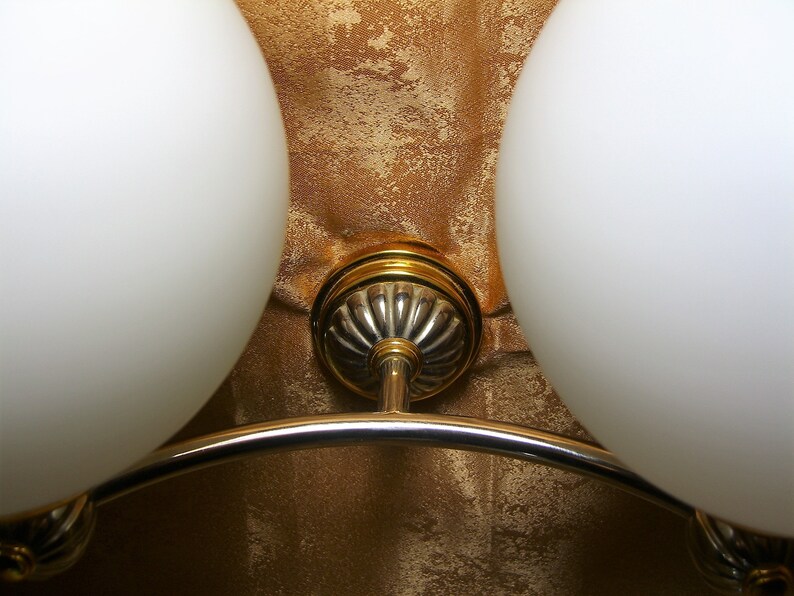 May include: A close-up of a gold and silver ceiling light fixture with two white glass globes. The fixture has a curved arm connecting the two globes.