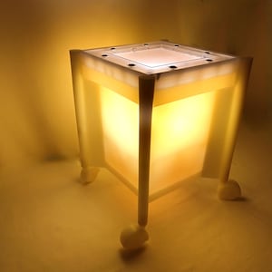 May include: A white, cube-shaped lamp with a clear top and a warm, yellow glow. The lamp is on wheels and has a modern, minimalist design.