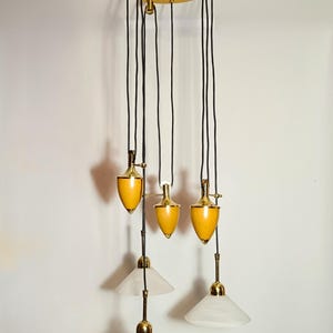 Könnte beinhalten: Eine sechsflammige Pendelleuchte mit einem Holzbaldachin und Messingakzenten. Die Leuchte hat fünf weiße Alabasterschirme und einen gelben Schirm.